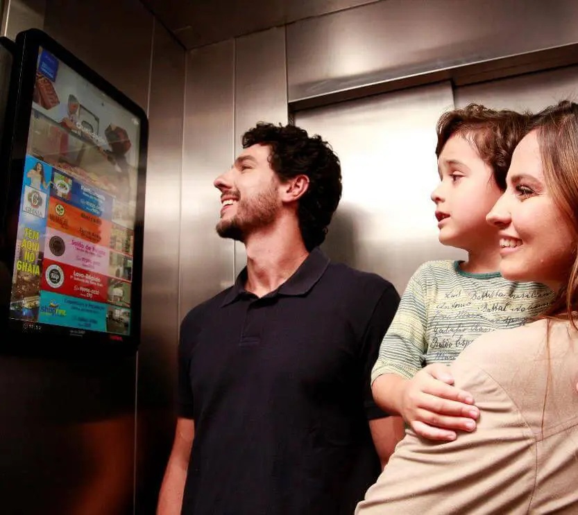 Familia composta por um homem, uma mulher e uma criança de colo dentro do elevador olhando um anúncio em uma tela